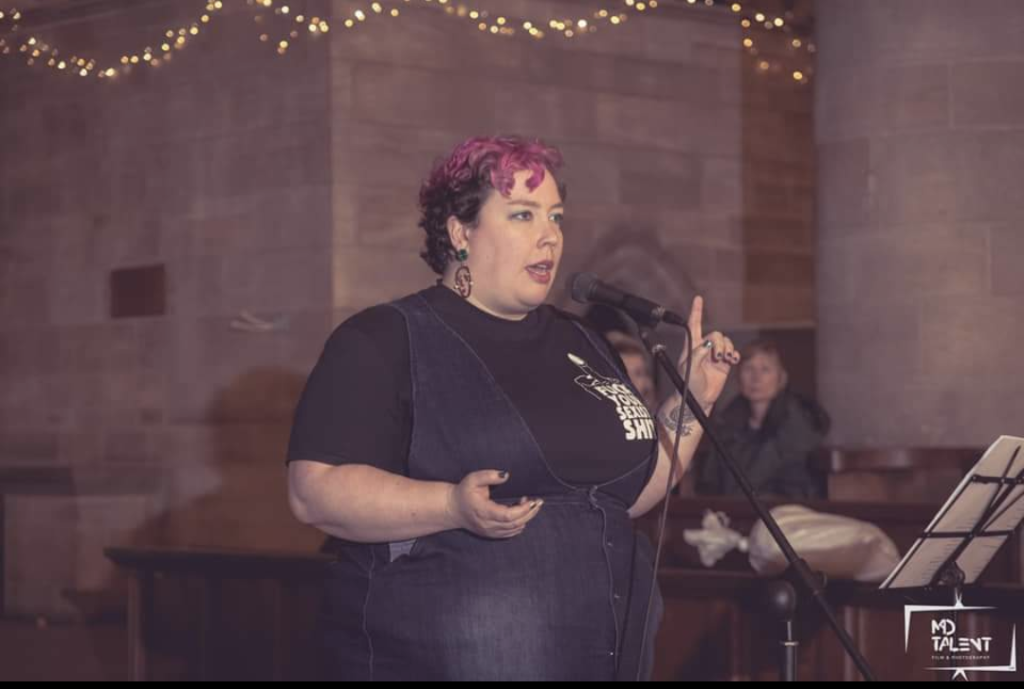 A performance shot of a fat, white person with short, curly, mid-brown hair dyed bright magenta on top is standing behind a microphone and music stand in what appears to be a large, old church. They are immaculately made-up with large hoop earrings and metallic nail polish, gesturing authortitatively, one finger raised, the other hand cupped beside them. They are wearing a dark, navy blue pinafore dress over a black teeshirt which bears the caption "Fuck Your Sexist Shit", and have a tattoo on the wrist of the hand pointing upwards.