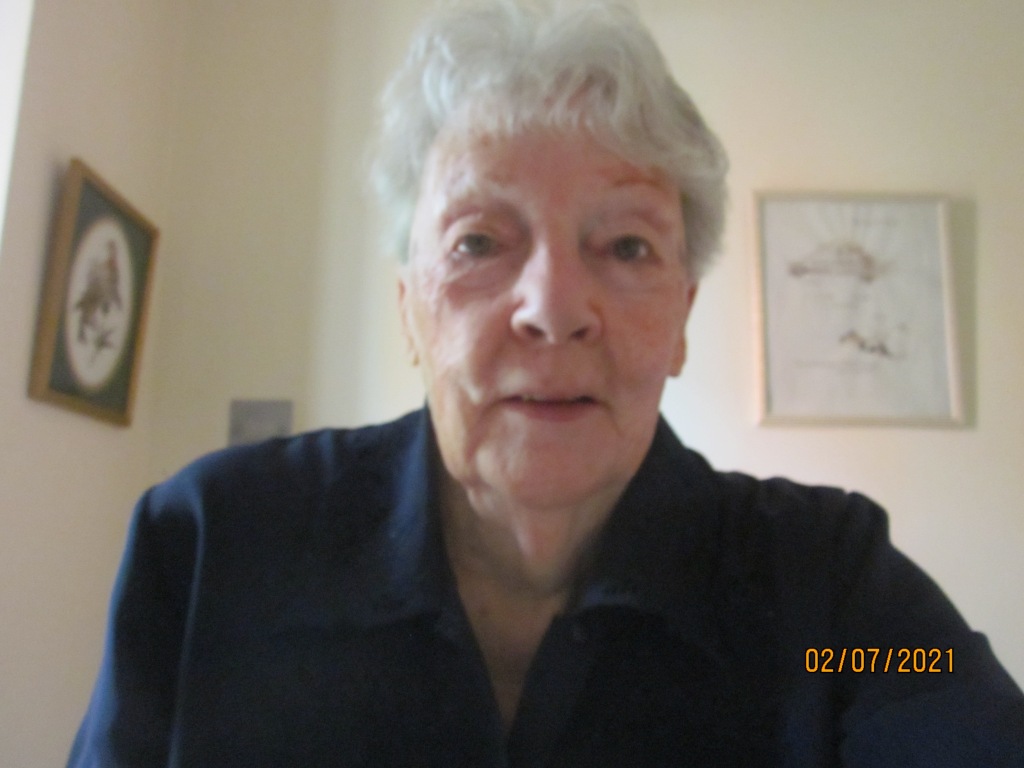 photo of an elderly, white woman with short, lightly curling silver hair, staring directly at the viewer with large, blue eyes. She is wearing a dark-blue, collared shirt and is smiling slightly, the cream, interior walls behind her showing a couple of framed pictures. At the bottom right-hand corner is the date 02/07/2021 in orange type.